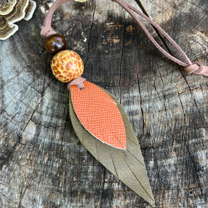 A handmade leather leaf bag charm with an orange textured center, layered over a brown leather leaf, featuring a patterned orange and white bead and a dark brown wooden bead on a rustic wood background.