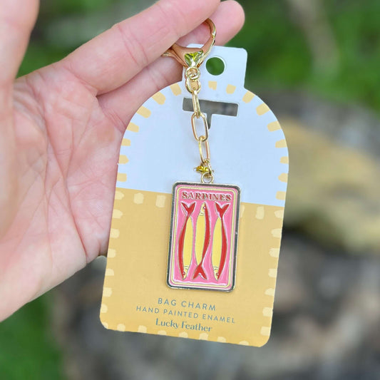 A hand holds a gold-tone keychain charm featuring a pink and yellow enamel sardine tin design, attached to a white and yellow retail display card labeled 'Sardines' and 'Bag Charm'.