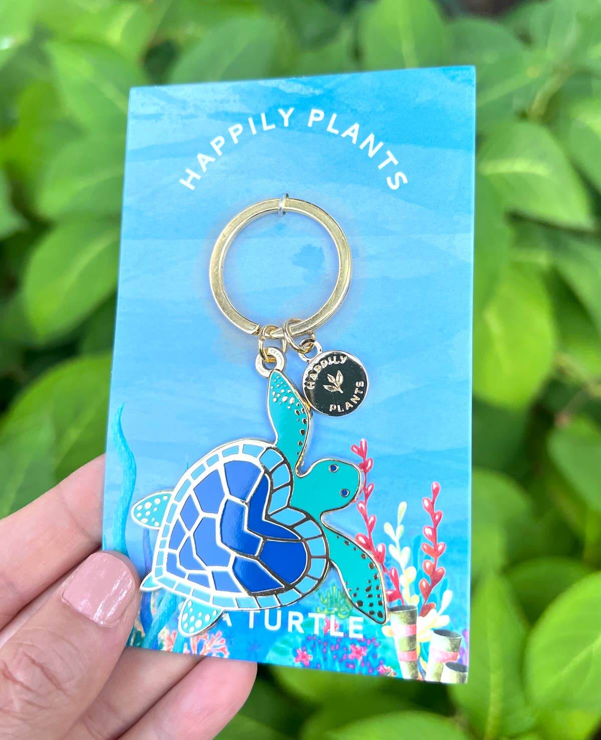 Turtle keychain on a blue card with 'Happily Plants' branding, held against a green leafy background.