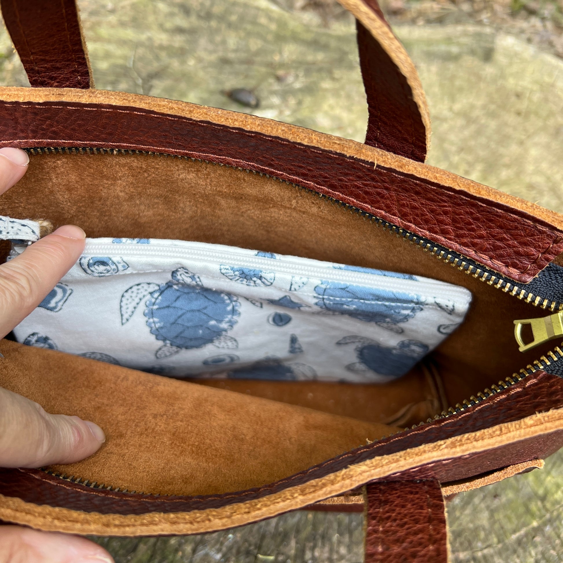 Brown leather bag with a white pouch featuring blue turtle designs inside, held by a hand.