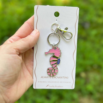 A hand holds a pink and silver enamel seahorse keychain on a white display card. The background is a soft-focus green outdoor setting.
