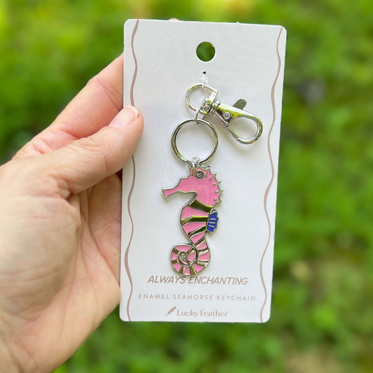 A hand holds a pink and silver enamel seahorse keychain on a white display card. The background is a soft-focus green outdoor setting.