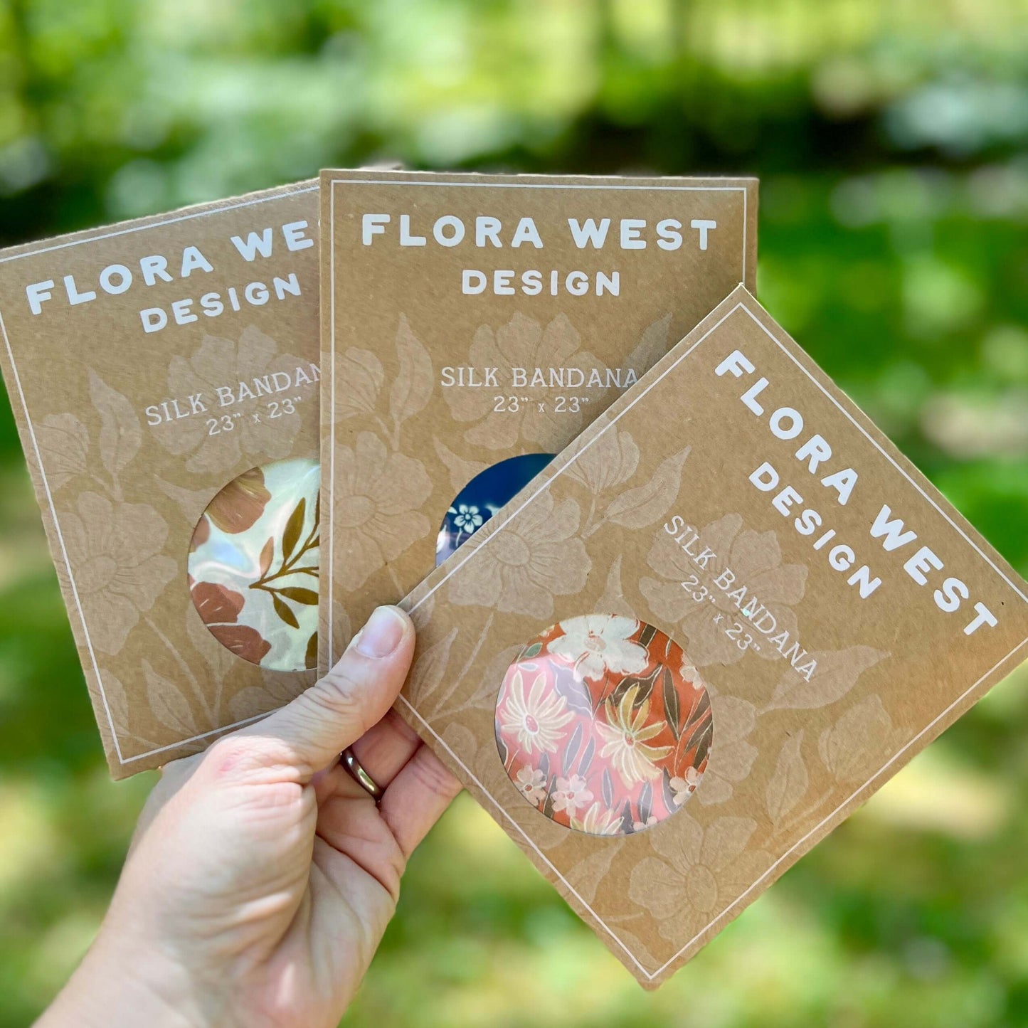 Three silk bandana packages from Flora West Design held by a hand with a blurred green background.