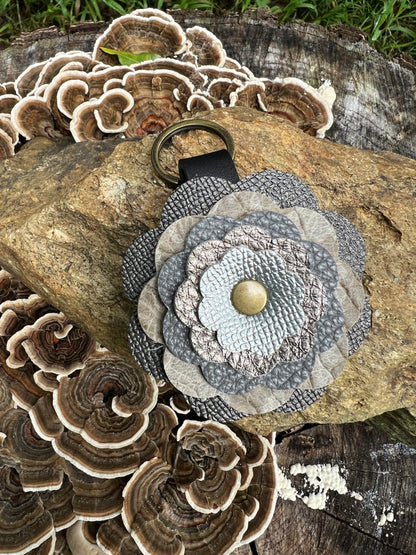 A leather flower-shaped keychain or bag charm with layered gray and beige patterned petals, a brass-tone center button, and a black strap with a metal ring, displayed on a log outdoors with mushrooms in the background.