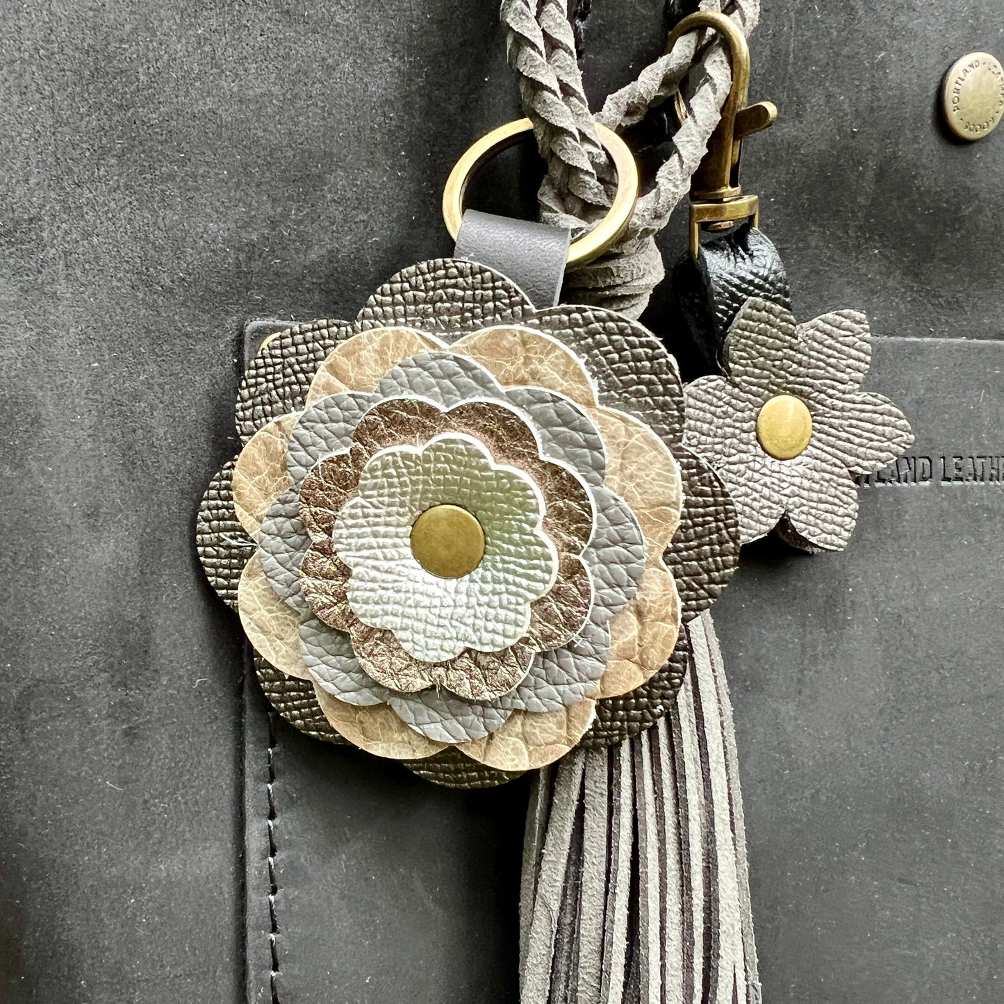 A close-up of a floral leather keychain or bag charm with layered flower petals in beige, taupe, gray, and off-white, gold-tone metal rings and snaps, and a long gray tassel, shown attached to a dark gray leather surface.