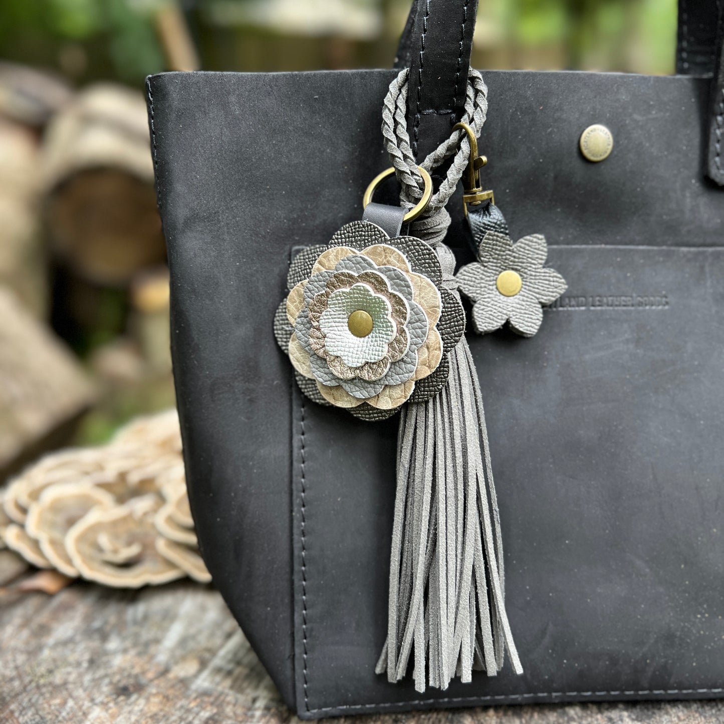 A close-up of a black leather tote bag featuring decorative keychain charms, including a braided strap, a large layered fabric flower in gray and beige tones, a smaller matching flower, and a gray tassel.