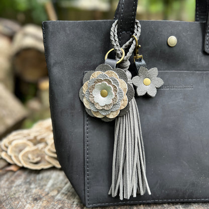 A close-up of a black leather tote bag featuring decorative keychain charms, including a braided strap, a large layered fabric flower in gray and beige tones, a smaller matching flower, and a gray tassel.