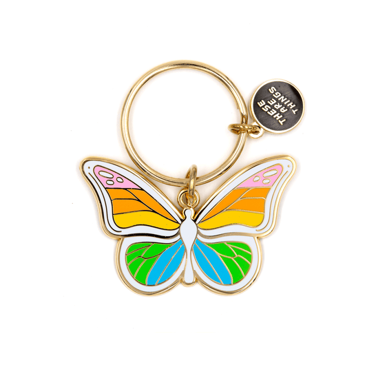 A butterfly-shaped keychain charm with a gold-tone metal ring and a small round tag that reads 'THREE THINGS'. The butterfly has multicolor enamel wings in pink, orange, yellow, blue, and green on a white background.