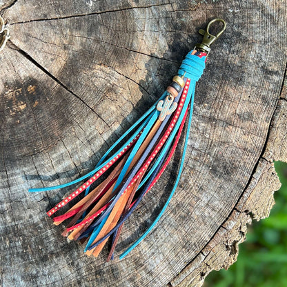 A colorful leather tassel keychain with multiple braided and flat leather strands in blue, red, orange, and brown, featuring a small wooden charm and a brass clasp, displayed on a rustic wood log background.
