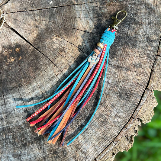 A colorful leather tassel keychain with multiple braided and flat leather strands in blue, red, orange, and brown, featuring a small wooden charm and a brass clasp, displayed on a rustic wood log background.