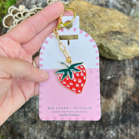 A hand holds a strawberry-shaped bag charm keychain on its pink retail card. The keychain has a gold-tone lobster clasp and chain, with a red enamel strawberry featuring green leaves and white seed details.