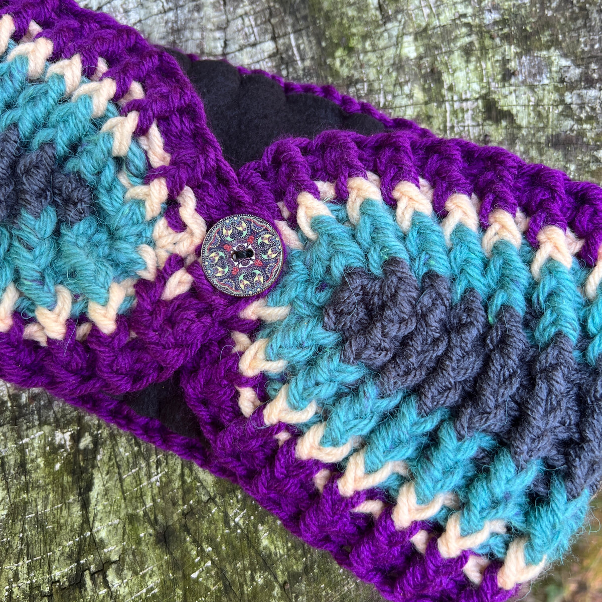 A close-up of a handmade crocheted headband featuring a vibrant purple, teal, and cream chevron pattern, secured with a decorative button, laid on a textured wooden surface.