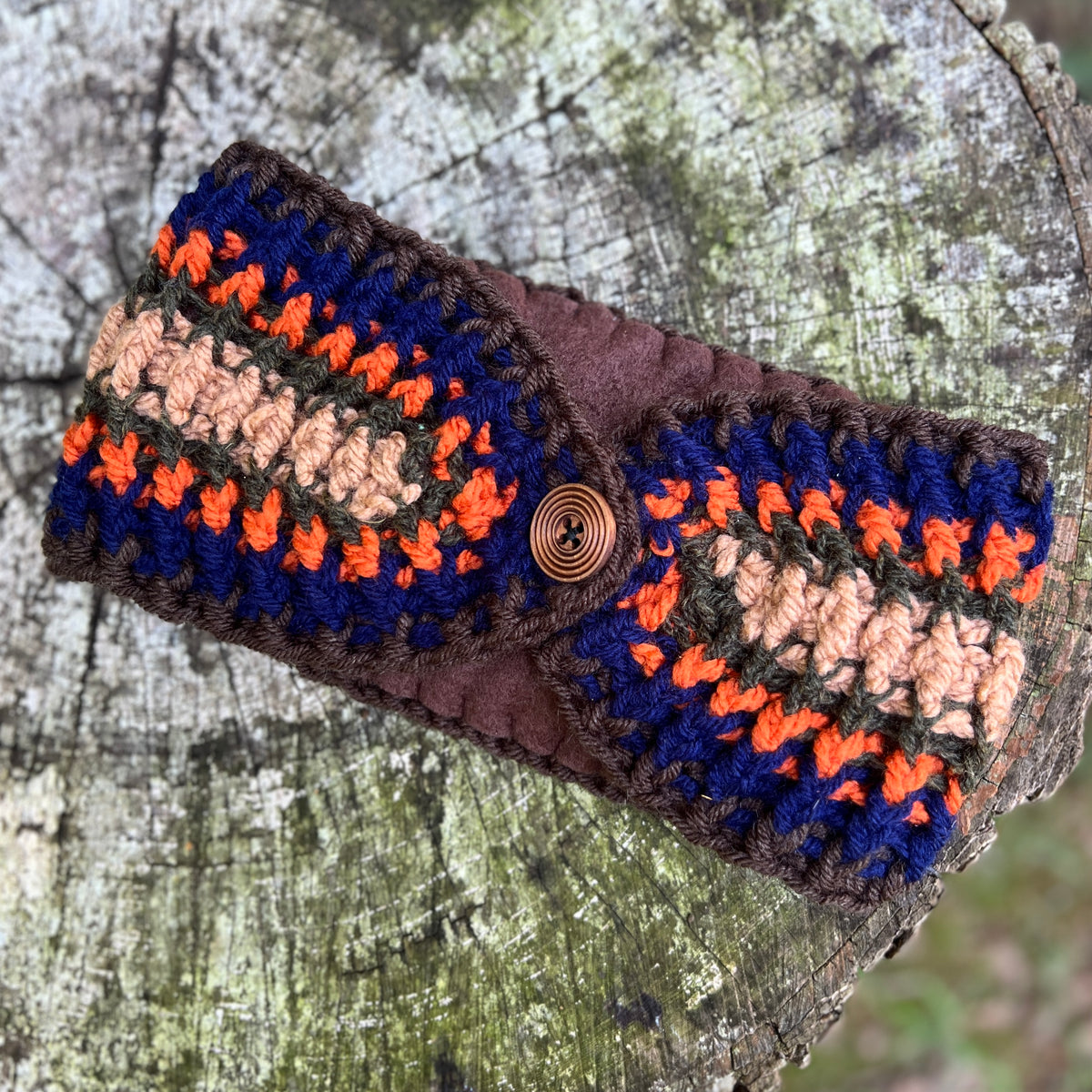 A handmade crocheted winter headband with a flap closure and a spiral button, resting on a weathered tree stump. The ear warmer features a multi color pattern of navy blue, orange, beige, and brown yarn.