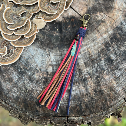 A handmade leather tassel keychain with red, navy, and brown straps, a small turquoise charm, and a brass clasp, displayed on a rustic wood log next to a mushroom.