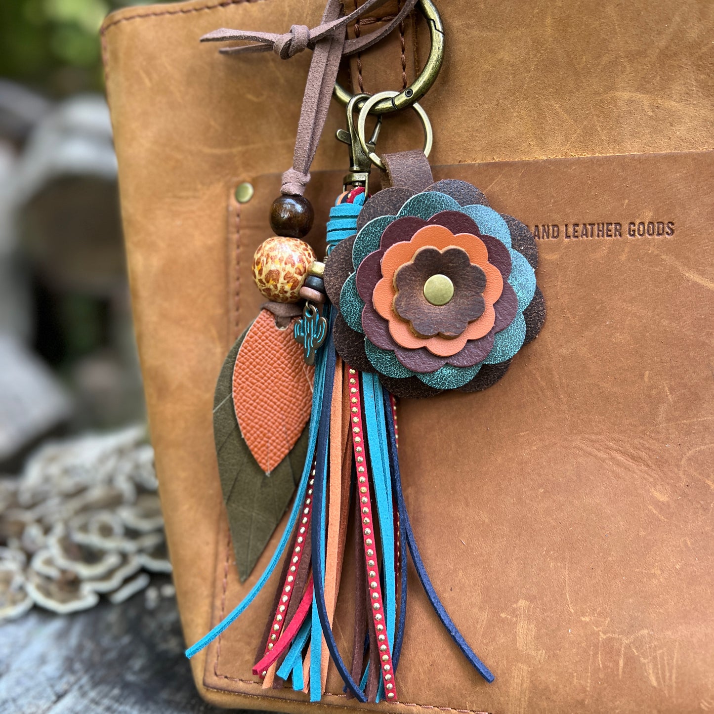 A bohemian-style leather keychain charm with layered flower petals, tassels, and decorative beads, attached to a brown leather bag. The bag has 'PORTLAND LEATHER GOODS' embossed on the side.