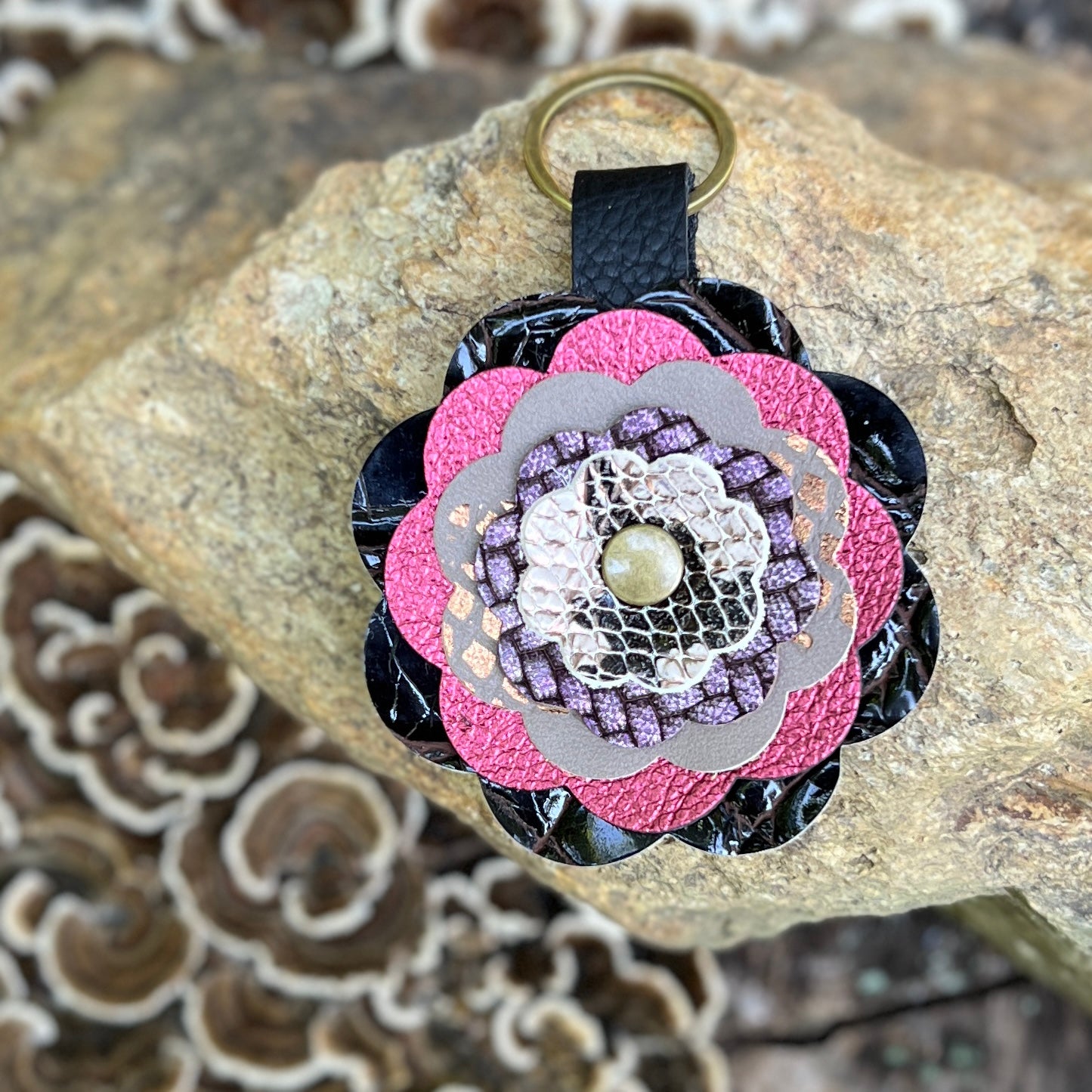 A handmade floral keychain or bag charm with layered pink, purple, and gray textured petals, a gold-tone metal ring, and a black strap, displayed on a natural rock.