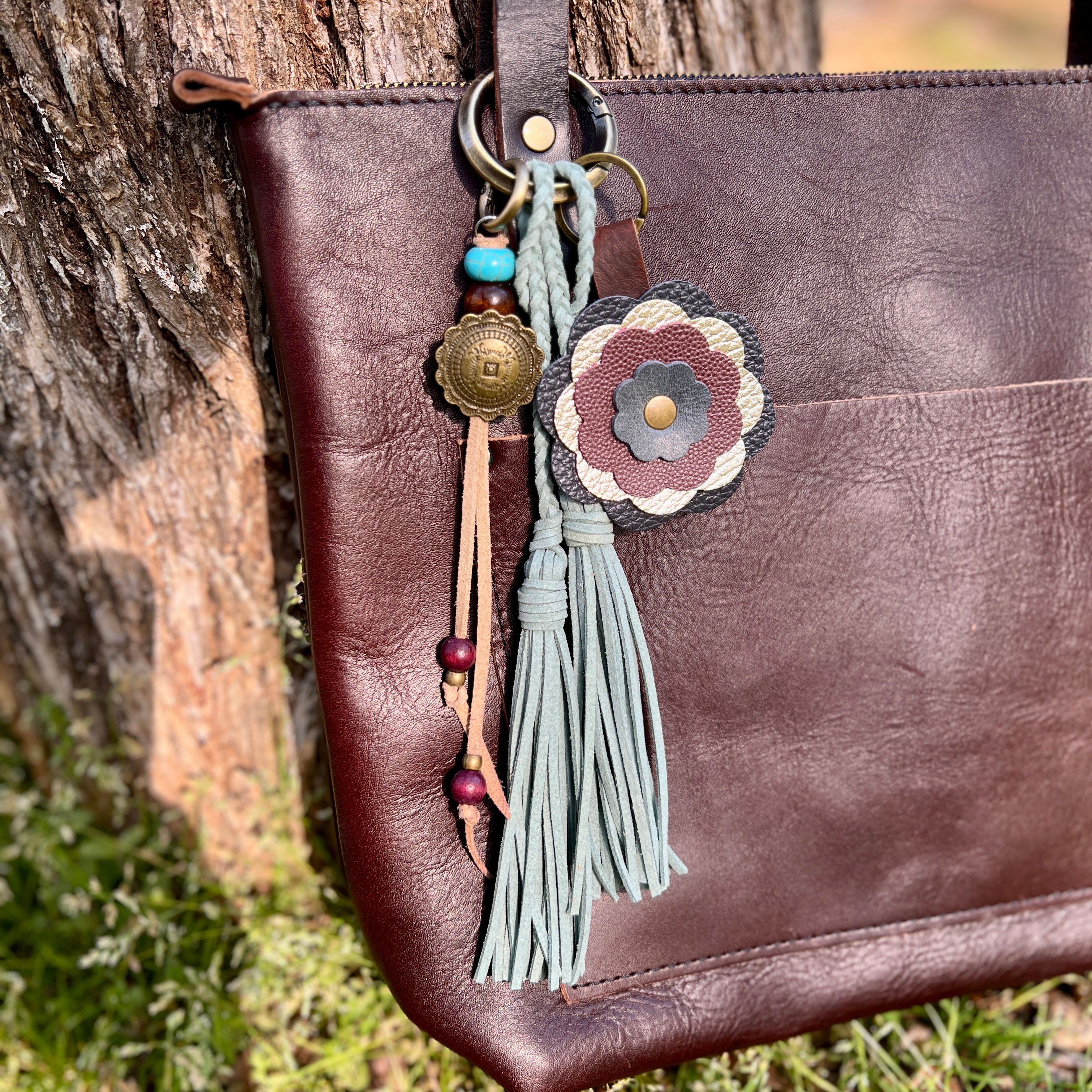 Brown leather bag with decorative keychain featuring a flower and tassel against a natural background.