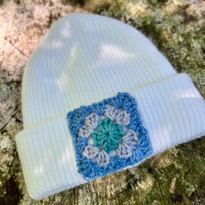 A white ribbed knit beanie hat with a crocheted granny square patch in blue, white, and green, resting on a textured tree trunk.