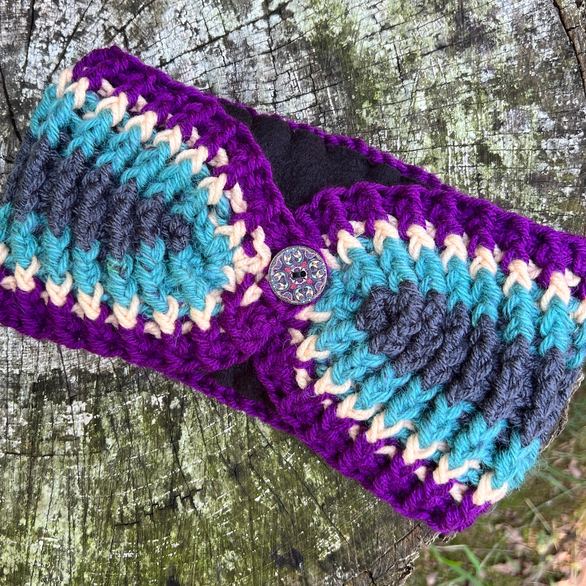 A handmade crocheted ear warmer or headband with a zigzag pattern in purple, teal, dark gray, and cream, featuring a decorative button closure, displayed on a weathered tree trunk.