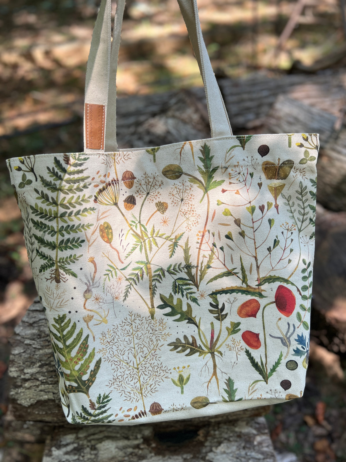 A canvas tote bag with a detailed botanical and bird illustration print, featuring green ferns, red berries, and small birds on an off-white background. The bag has light beige shoulder straps with a small brown leather accent and is photographed outdoors on a rustic stone surface with a blurred forest background.