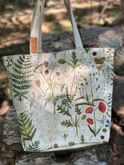 A canvas tote bag with a detailed botanical and bird illustration print, featuring green ferns, red berries, and small birds on an off-white background. The bag has light beige shoulder straps with a small brown leather accent and is photographed outdoors on a rustic stone surface with a blurred forest background.