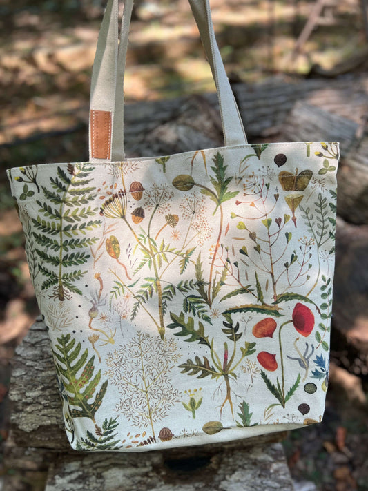 A canvas tote bag with a detailed botanical and bird illustration print, featuring green ferns, red berries, and small birds on an off-white background. The bag has light beige shoulder straps with a small brown leather accent and is photographed outdoors on a rustic stone surface with a blurred forest background.