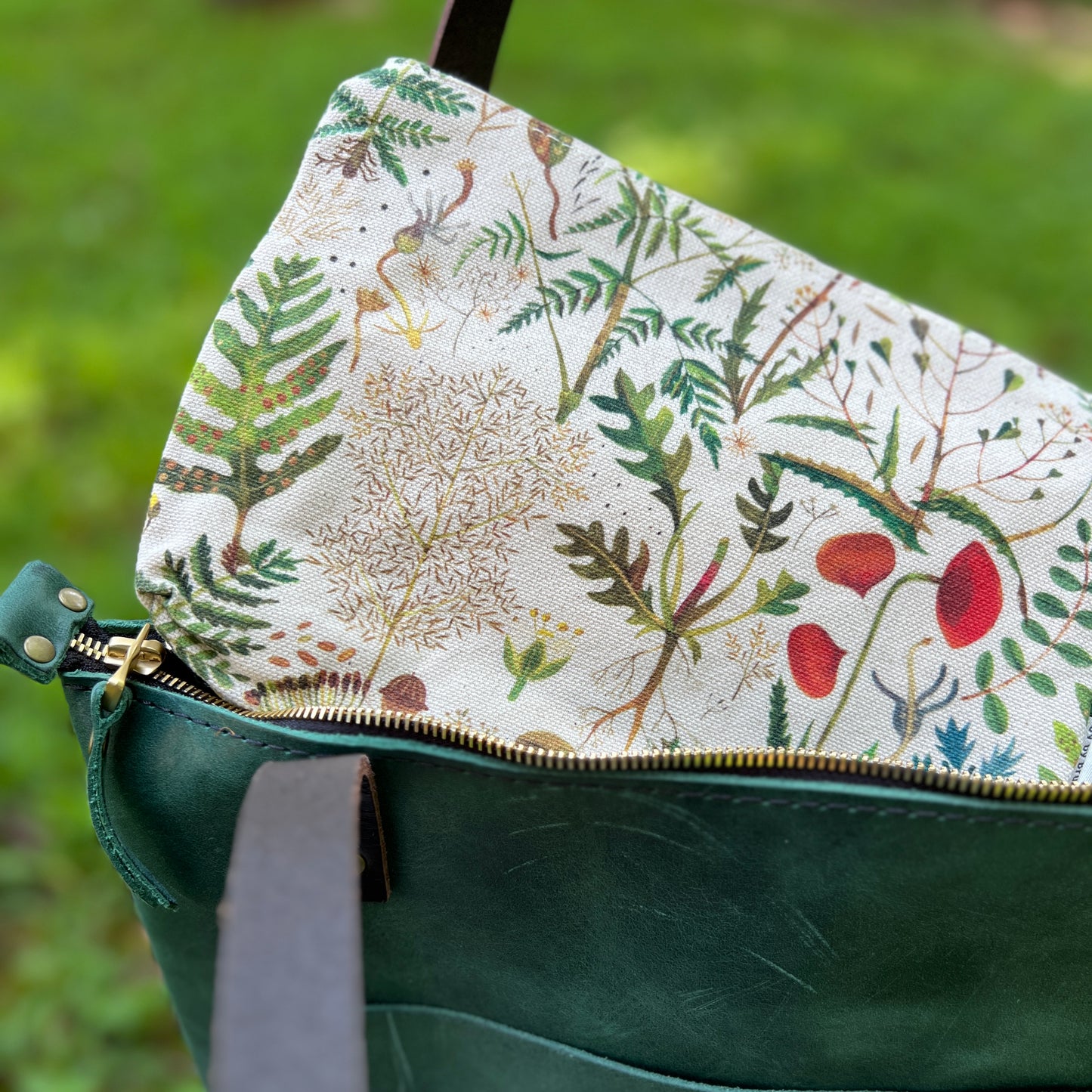 Close-up of a green leather bag with a gold zipper and a cream fabric lining printed with botanical illustrations of leaves, flowers, and birds, set against a blurred green grass background.