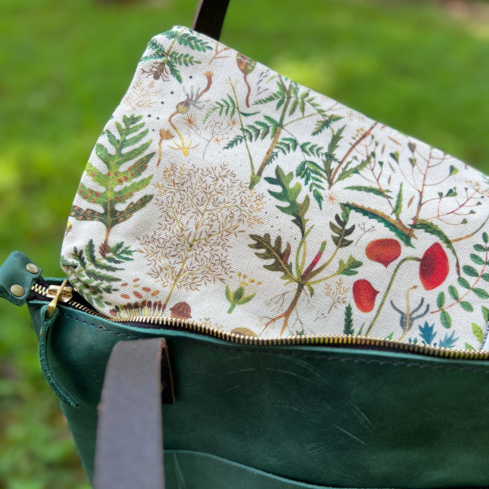 Close-up of a green leather bag with a gold zipper and a cream fabric lining printed with botanical illustrations of leaves, flowers, and birds, set against a blurred green grass background.
