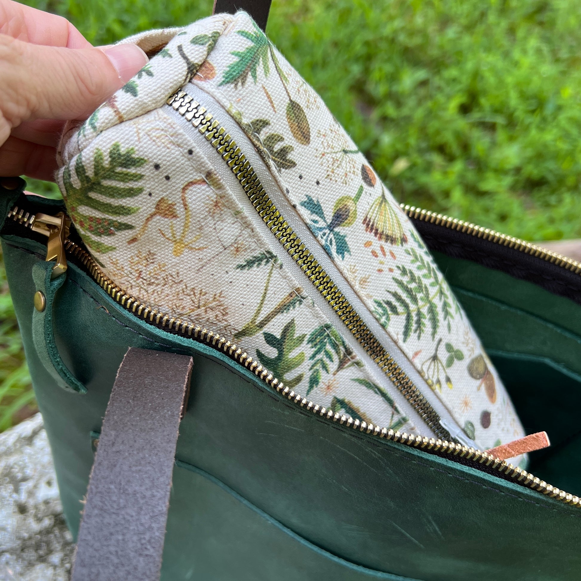 A close-up of a hand holding a small fabric pouch with a botanical print pattern, shown inside a dark green leather bag with gold-toned zippers.