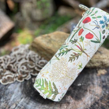 A cylindrical fabric pouch with a natural canvas background and a botanical print of green leaves, red berries, and blue flowers, resting on a rustic wooden surface with blurred rocks in the background.