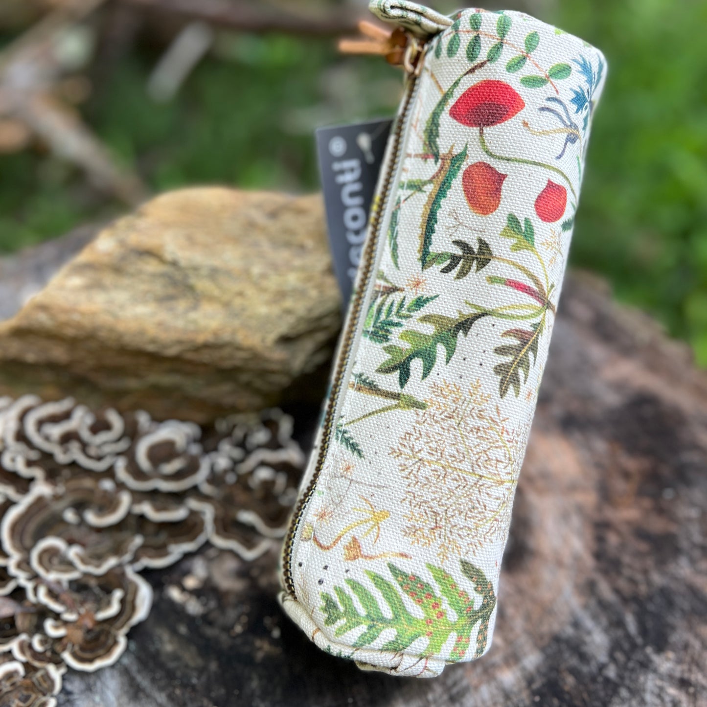 A fabric zippered pencil case with a botanical print of leaves, flowers, and mushrooms on a cream background, resting on a tree stump outdoors.