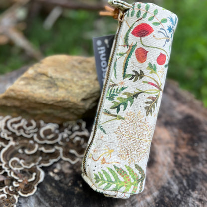 A fabric zippered pencil case with a botanical print of leaves, flowers, and mushrooms on a cream background, resting on a tree stump outdoors.