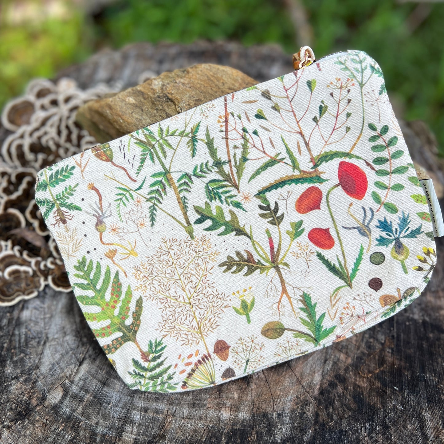 A small zip pouch with a white background and a detailed botanical print of green leaves, red poppies, and yellow flowers, resting on a rustic wooden surface.