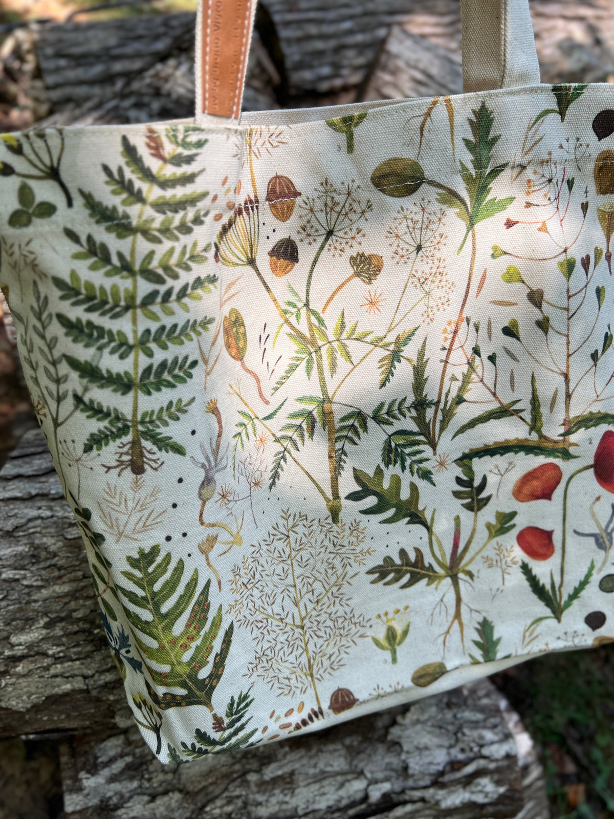 A canvas tote bag with a detailed botanical print of ferns, wildflowers, and acorns, featuring light brown leather handles, resting on a rustic log outdoors.