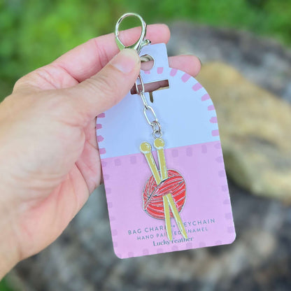 A hand holds a keychain with a silver lobster clasp, featuring a red and orange striped ball of yarn and gold knitting needles. The keychain is attached to a light purple and pink display card labeled 'Lucky feather'.